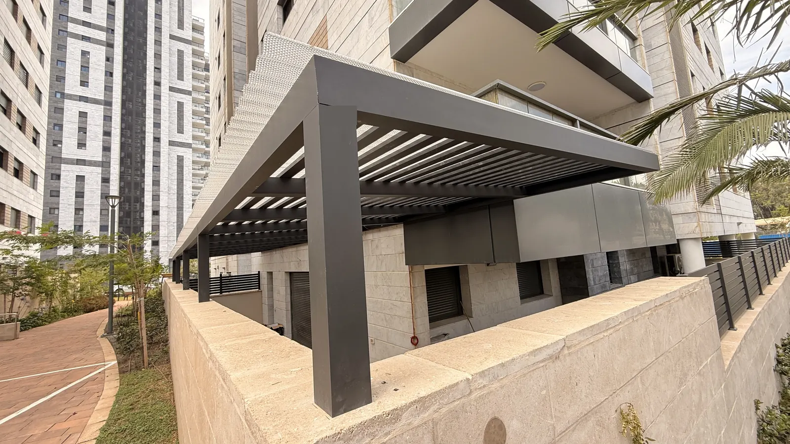 מצללת אלומיניום אפורה עם רפפות מעל מרפסת גן על קיר אבן, בבניין מגורים בראשון לציון